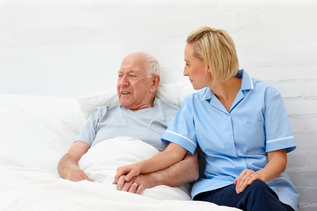 A dedicated nurse sitting attentively beside an elderly man in bed, representing CareHubSupport's round-the-clock live-in care service