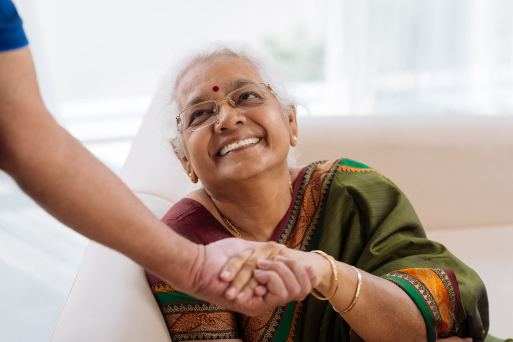 A South Asian elderly woman smiling warmly as a carer holds her hand, reflecting the trusted specialist support CareHubSupport provides